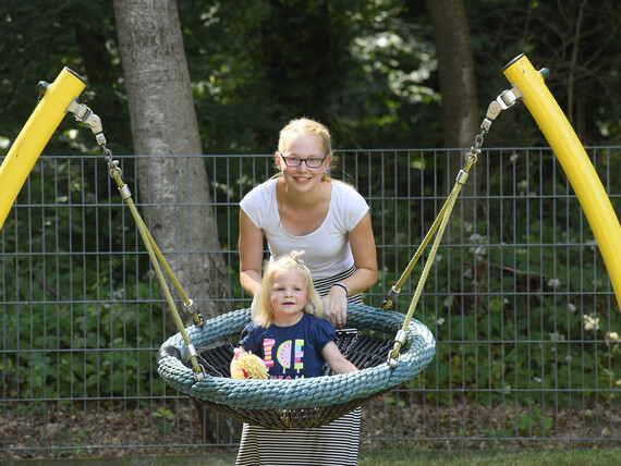 Ein Kind sitzt zufrieden in der Nestschaukel, eine lächelnde Erzieherin im Hintergrund.