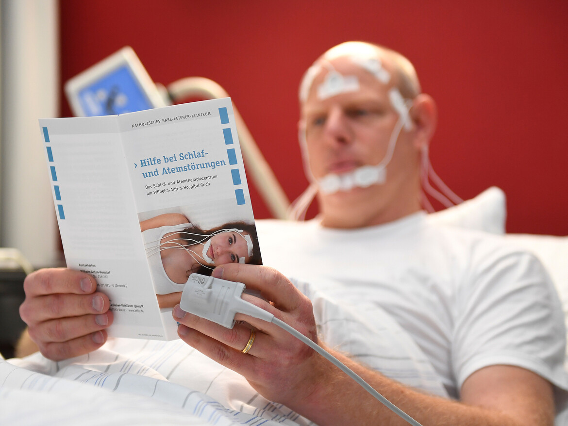 Ein verkabelter Mann liegt im Bett und liest einen Flyer des Schlaflabors.