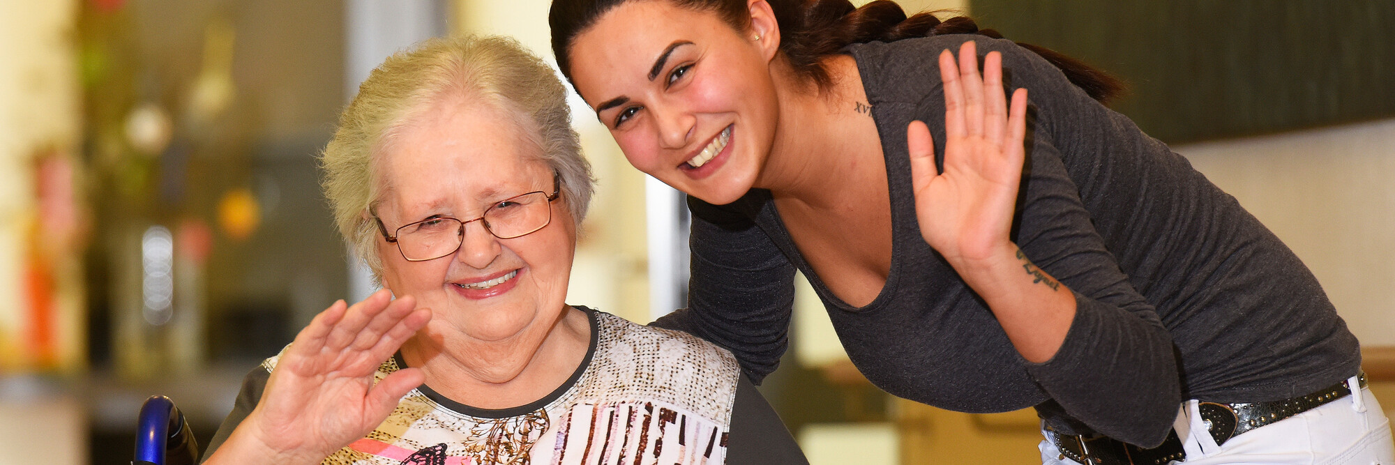 Eine Seniorin und eine Altenpflegerin winken gemeinsam in die Kamera.