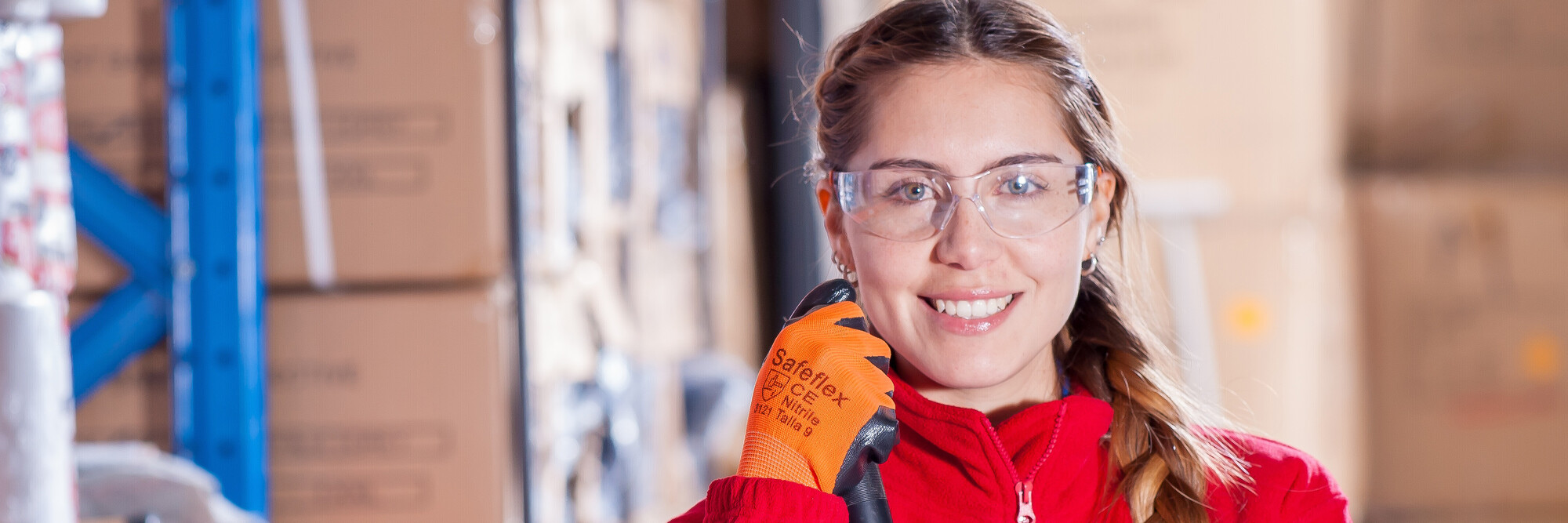 Eine junge Lagerarbeiterin mit Schutzbrille und Handschuhen blickt in die Kamera.