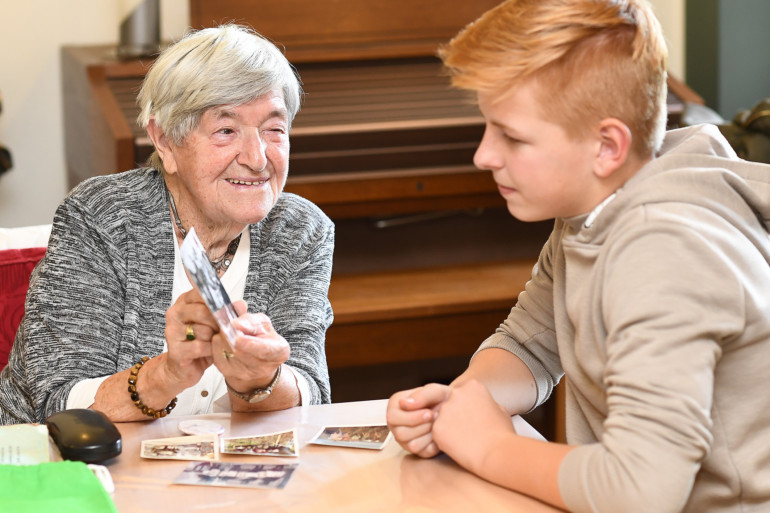 Eine Seniorin zeigt einem Jugendlichen Fotos aus ihrer Kindheit - beide lächeln.